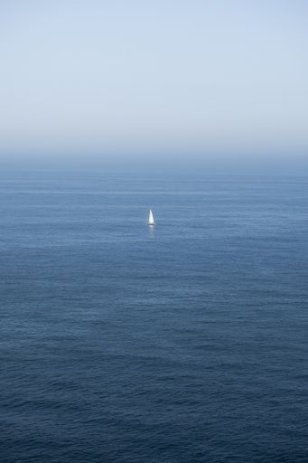 An aerial shot of the endless ocean with a white sailboat under a cloudy sky