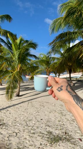 A closeup of a male hand with a tattoo holding a cup of coffee on the beach
