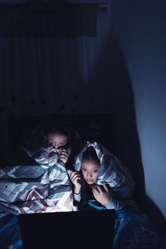 A vertical shot of two Hispanic Young women at a sleepover watching movies at dark on a laptop