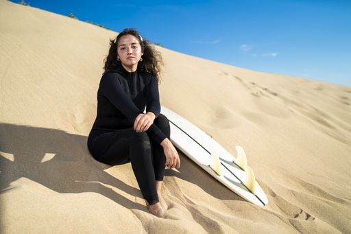 A shallow focus shot of an attractive female sitting on a sandy hill with a surfboard on the side