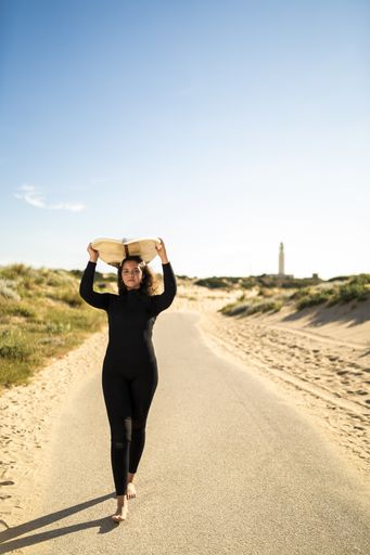 A vertical shot of an attractive female carrying a surfboard above her head while walking in the middle of the road in Spain