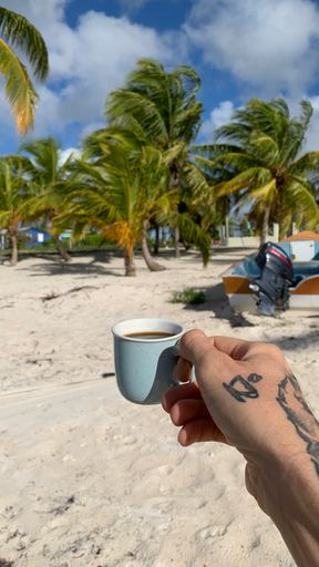 A closeup of a male hand with a tattoo holding a cup of coffee on the beach