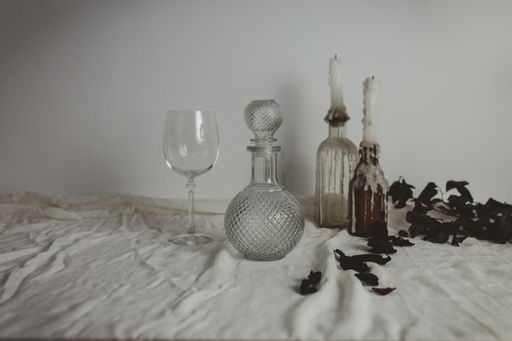 A vase, bottles, glass and candles on a white surface next to dry flower petals and leaves
