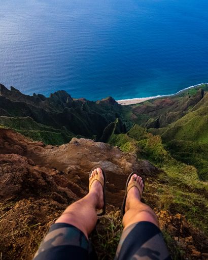 A vertical high angle shot of a person sitting on a cliff and looking down on the Napali Coast, Kauai, Hawaii