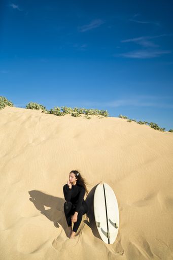 A vertical shot of an attractive female sitting on a sandy hill with a surfboard on the side