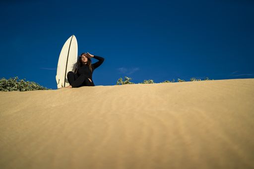A low angle shot of an attractive female sitting on a sandy hill with a surfboard on the side