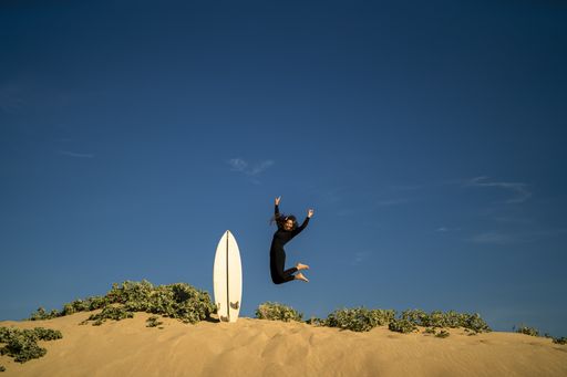 An attractive female jumping on a sandy hill with a surfboard on the side