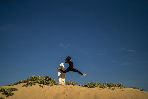 A female jumping on a sandy hill with a surfboard at the back
