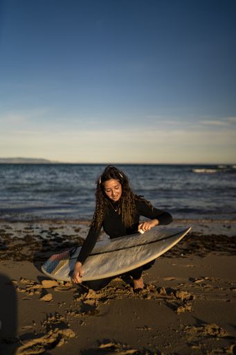 A vertical shot of a smiling female waxing her surfboard at the beach in Spain