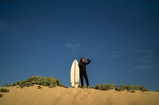 An attractive female posing on a sandy hill with a surfboard on the side