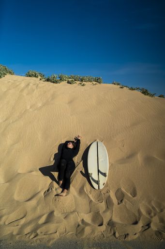 A vertical shot of an attractive female lying down on a sandy hill with a surfboard on the side