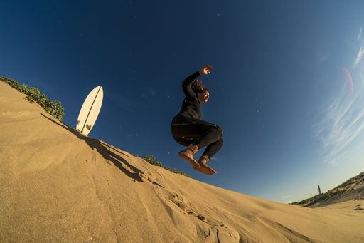 A low angle shot of a female jumping on a sandy hill with a surfboard on the side