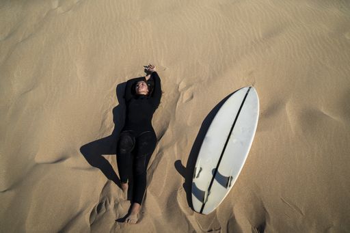 An aerial shot of an attractive female lying down on a sandy ground with a surfboard on the side