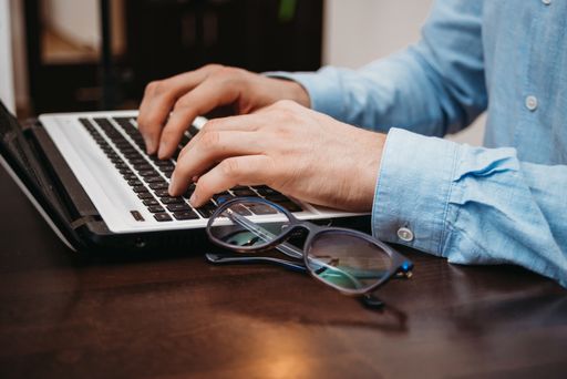 The male hands typing a text on the laptop on the table near a pair of glasses in the office