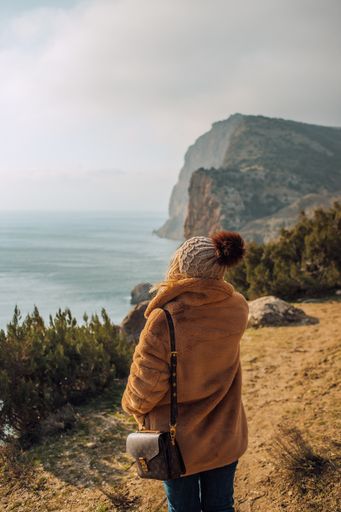 A vertical, back view of  a girl with a brown fur coat walking along a path near the sea