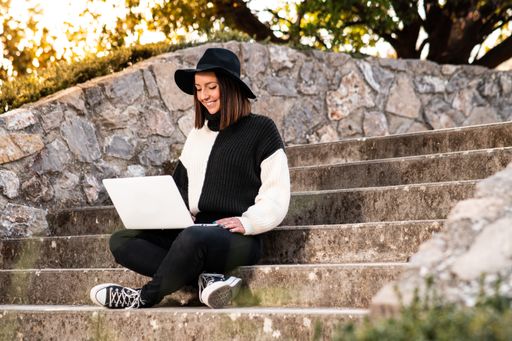 A cute cheerful Caucasian woman in a hat
sitting on the stairs in a park working on her computer