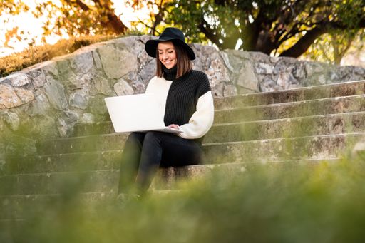 A cute cheerful Caucasian woman in a hat
sitting on the stairs in a park working on her computer