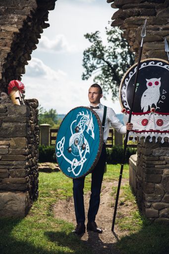 A vertical shot of a man in a suit with a shield and a spear in his hands posing at an ancient castle