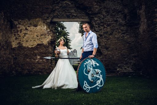 Beautiful couple in suit and wedding dress poses in front of camera with spear and shield in ancient castle