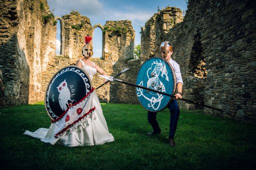 Beautiful couple in suit and wedding dress poses in front of camera with spear and shield in an ancient castle