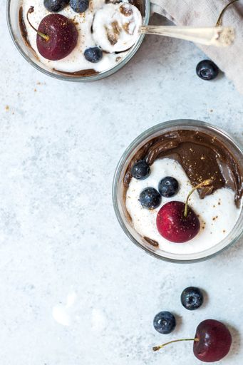 An overhead vertical shot of chocolate avocado puddings with cherries on top on a white surface