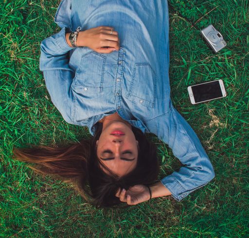An overhead shot of a young female relaxing on the grass with her phone and camera on the side