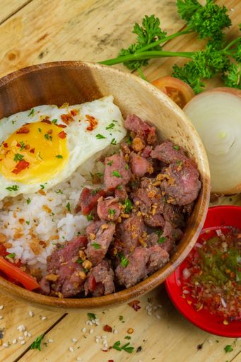 A dish with meat, rice and omelet served in a wooden bowl and some vegetables nearby