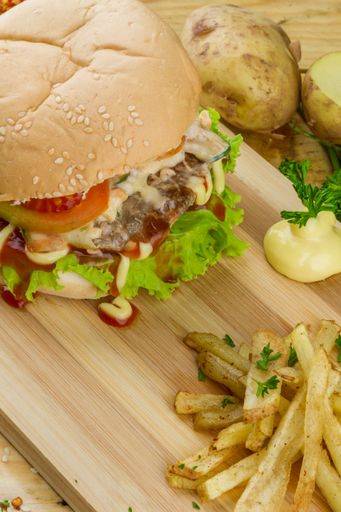A big burger with some French fries and souse on a wooden tray and potatoes nearby