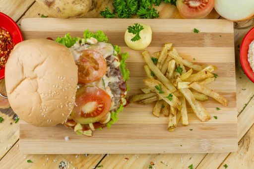 A burger and French fries with some souse on a wooden tray and vegetables nearby