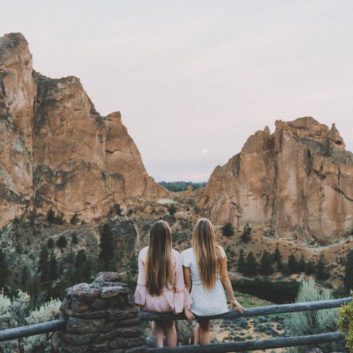 A beautiful shot of two females sitting on a wooden fence enjoying the view with mountains and a cloudy sky in the background