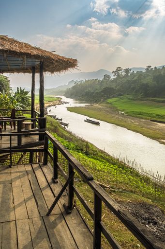 A view of a hut with a wooden floor by a river, grass against a blue cloudy sky in Thanchi, Bangladesh