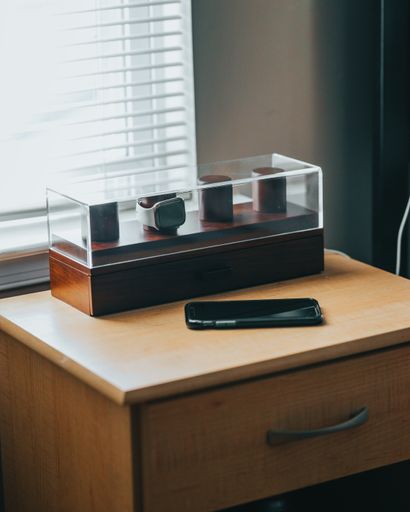 A vertical shot of a watch in a glass box and a phone on a wooden surface
