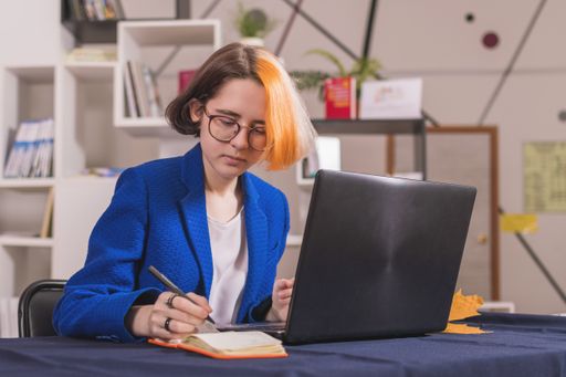 Young modern girl with multi-colored hair in blue business suit sits at table with laptop. Makes notes in an orange notebook.
