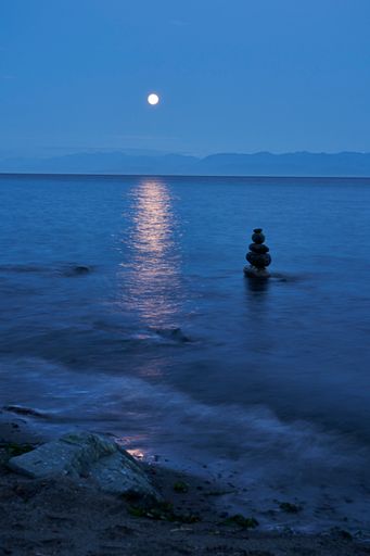 Evening scene of an inuksuk in a blue ocean with the moon casting its reflection on the water. Glimpses of a large rock on the beach.