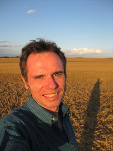 A vertical shot of a male smiling and looking at the camera standing in a grassy field with a blurred  background