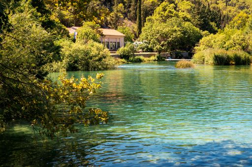 A beautiful view of a calm river with a house on the shoreline surrounded by green trees on a sunny day