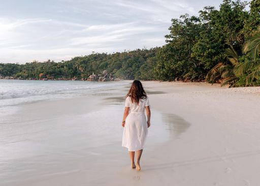 A Rear view of young woman wearing white summer dress, standing on Anse Lazio beach on Seychelles