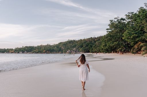A Rear view of young woman wearing white summer dress, standing on Anse Lazio beach on Seychelles