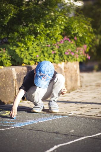 A vertical shot of a boy with a blue hat drawing with blue chalk outdoors