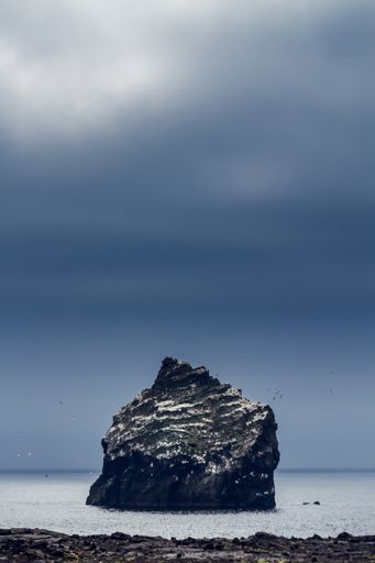 A vertical shot of the cloudy sky over the sea with a huge rock in it in Icelandic area