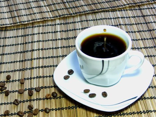 A closeup of a cup of black coffee with coffee beans spilled around on a bamboo fence roll