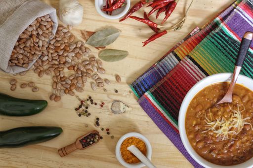 A top view of bowl of chili with pinto beans on colorful carpet near the peppers, spices and dry beans