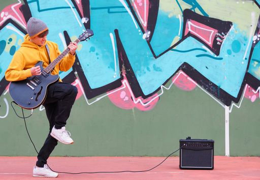 A young boy playing guitar in the street, in front of a wall with graffiti