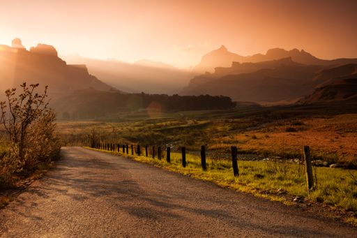 A concrete road with a field on the side and a silhouette of mountains in the background during sunset