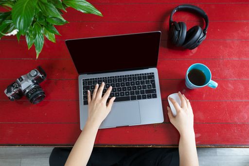 A top view of female hands on a modern laptop with a camera, green plant, headphones, and a cup of coffee