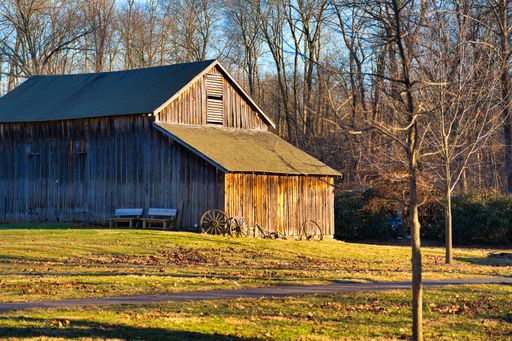A beautiful shot of a red wooden barn in the grassland with trees against a blue sunny sky