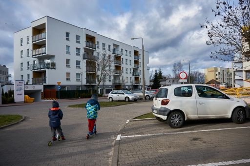 A close-up shot of two kids with scooters on a street with parked cars and apartment buildings