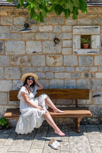 A Beautiful happy young woman wearing white dress, sitting on bench in front of old stone house