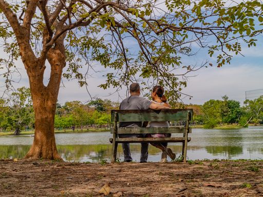 A couple sitting on a slanted park bench in a beautiful scenic park with a lake in front of them in Bangkok, Thailand.