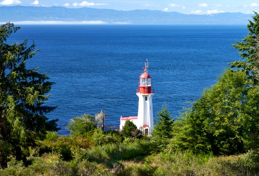 White and red lighthouse famed by coniferous trees with slightly rippled very blue ocean and  mountains in the background.